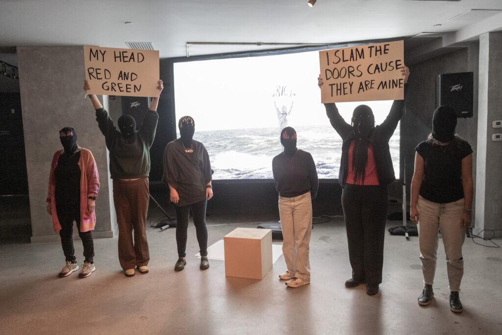 Six human figures, their figures covered in black hoods, stand in a line. Two of them hold white poster board signs above their heads, each with a phrase written in block letters with black marker. One reads "MY HEAD RED AND GREEN". The other says "I SLAM THE DOORS CAUSE THEY ARE MINE". Behind them is a video projection of a figure in a zebra-patterned Zentai suit, standing in a turbulent sea  and holding up a sign with the word "RIOT" printed on it.