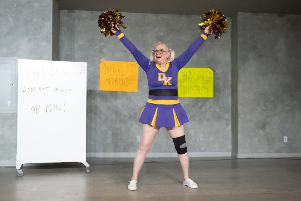 Tanya Mars stands, legs apart, arms held high, with glittery gold and purple pom poms in each hand. She is dressed in a purple cheerleader outfit with yellow accents, the letters DK on her chest. She wears white tennis shoes with a black and grey brace on one knee. Her blonde hair is separated into two pigtails by pink ribbons. She wears glasses and her mouth is open in a cheer, revealing a wide set of upper teeth. Scrawled on a whiteboard behind her is the phrase, "WHAT DO YOU GOT? HOW HOT IS HOT OH WOW!" Two large pieces of poster board are taped to the wall behind her. An orange one reads "SKEPTICALLY OPTIMISTIC" while a yellow one reads "OPTIMISTICALLY SKEPTICAL" Both also have numbers, some of them circled.