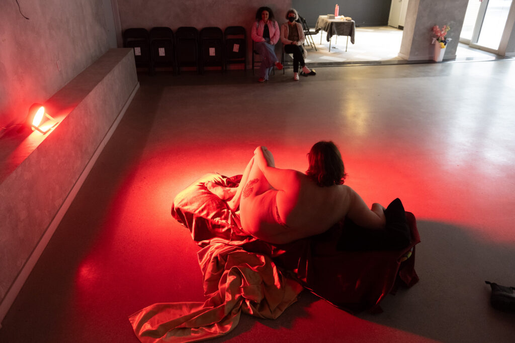 Tess Martens, shot from above and behind so that we only see her from the back, sits naked, curled up with her arm over one knee, facing two audience members sitting on folding chairs. She sits on satin-y shimmering fabrics, lit by a red light. Her brown hair is shoulder length, her figure appears fulsome, and a large tattoo is evident on one thigh.