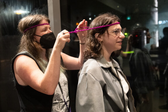 Heidi Nagtegaal ties a headband around an audience member's head. The artist has brown hair that falls down below her shoulders. She wears a braided red headband, a black face mask, and a sleeveless top. The audience member, a brown-haired woman in glasses and a grey zippered jacket over a white top, has an amused expression on her face as Heidi begins to tie together a thin, braided bright purple headband behind her.