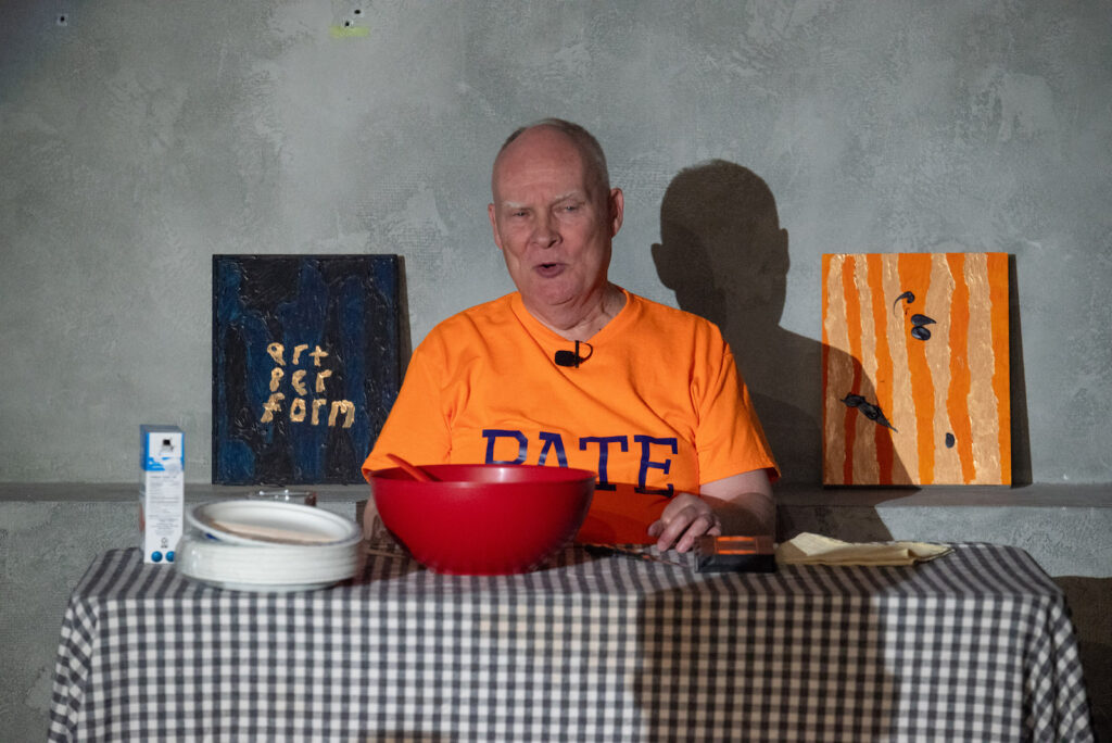 Andrew Paterson, an older man with receding hairline and white eybrows, sits behind a table that has been covered with a gingham tablecloth. He wears an orange t-shirt with the word PATE in blue letters on the front. Placed on the table are a box of salt, a stack of paper plates, a water glass, a red bowl with the handle of a red implement visible, a black videocassette, and a floded piece of cloth. he is flanked by two paintings. One painting has the words "art per form" in gold against a blue and black background. The other has several black blobs, like large paint drips, against a background of rough vertical strips of orange and gold.