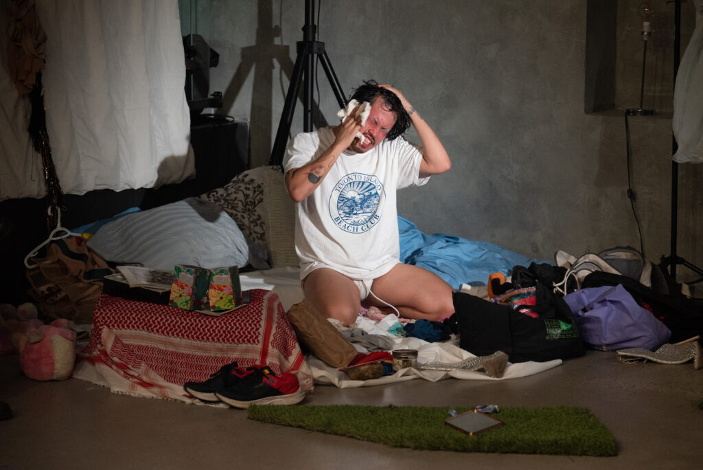 Tyler J. Sloane sits on their haunches on the floor, surrounded by clothes, fabric, bags and other objects. They wear a loose white t-shirt and white underwear. Their hands are lifted up to their head. One hand rubs their cheek with a white cloth, while the other rests on top of the head, tilting it to one side. their eyes are closed, their face is red, and their teeth are showing in a grimace. There are prominent tattoos on one wrist and the opposite forearm.