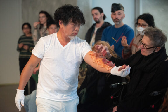 Daisuke Takeya, with black hair and a white goatee, is dressed in a white t-shirt, white pants, and short white gloves, stands parallel to the audience, one arm outstretched. He has several large pieces of salmon and tuna sashimi laid out along his arm and held in his palm. An audience member sitting on a walker, dressed in black with thick glasses, is selecting a piece of tuna from his arm. Other audience members, some holding pieces of sashimi, watch the action.