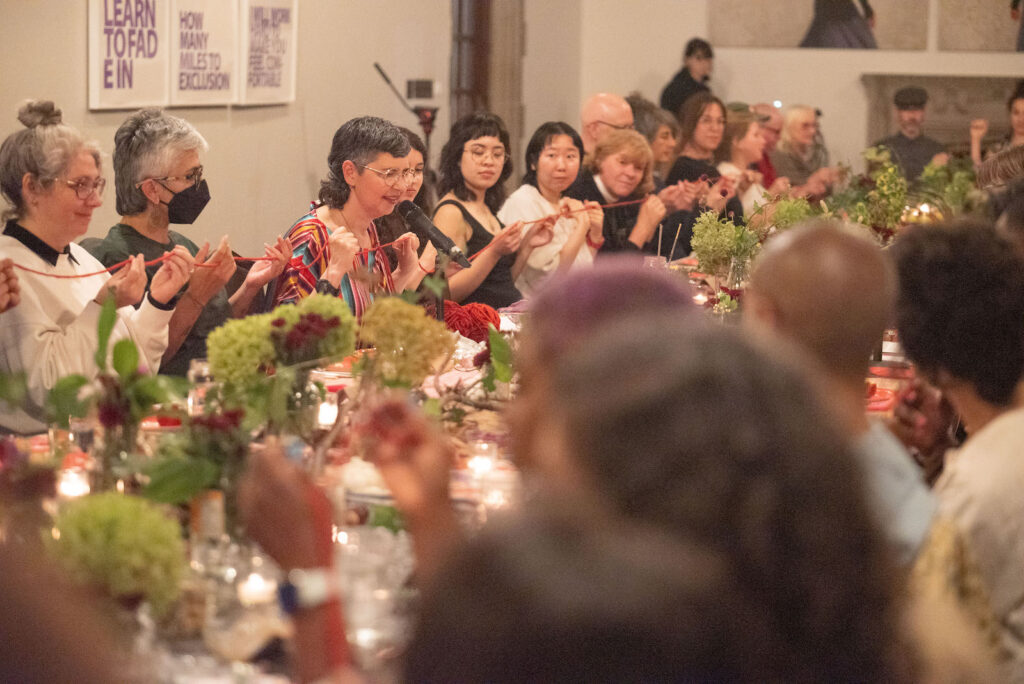 Many audience members sit around a long table festooned with flower arrangements. Their arms and hands are raised forward, their hands loosely holding a piece of red yarn that threads around the table. Some are looking at the yarn, some are looking at Jess Dobkin, who sits at the table speaking into a microphone. She wears a colourful striped dress, sliver-framed glasses, and a black wristwatch. She clutches a length of the yarn between her two fists.