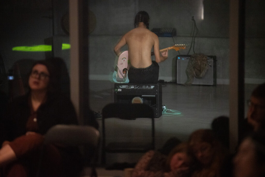 seth cardinal dodginghorse sits on a black amplifier with their back to the camera. They have dark hair with two braids, they are shirtless, and they are playing a pink electric guitar. They sit facing a second amplifier with a shirt draped over the front of it. The image is a reflection in the  window; in soft focus are the faces of several audience members, facing toward the camera but watching the performer.