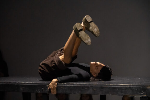 The artist lies on their back on a black riser, legs raised over their head. The heads of some audience members can be seen below the riser, looking forward. The artist has black hair, short in the front but long in the back, brown skin, with one arm spread out on the floor, hand gripping the edge of the riser, silver bands one the three middle fingers. They wear a long sleeve black top and brown pants rolled up above their knees. They wear bronze shoes.