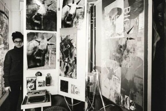 In this black and white photograph, a woman with glasses stands in a long, narrow room. She stands to the side of a large hinged display panel, taller than the woman and featuring multiple images. A manual typewriter rests on a small table in front. Other artworks can be seen on the side walls of the room.