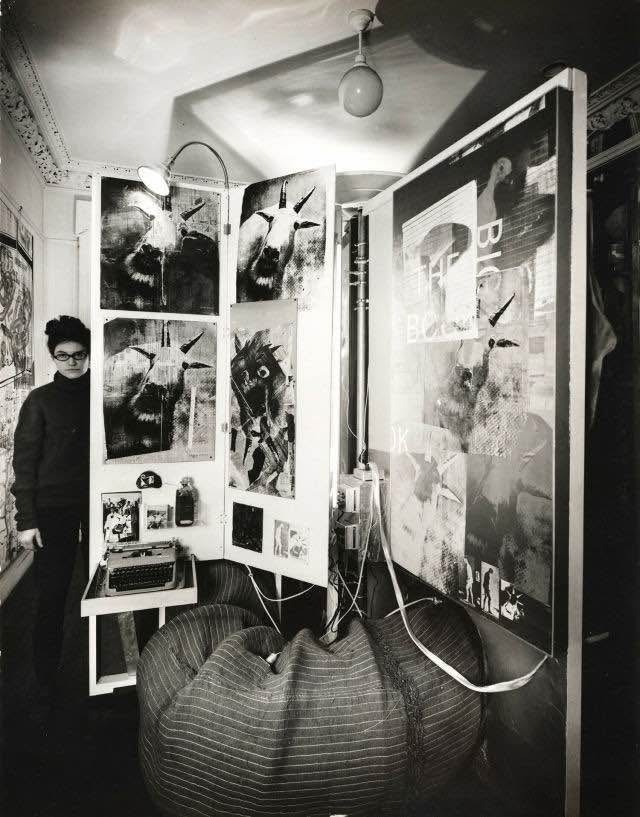 In this black and white photograph, a woman with glasses stands in a long, narrow room. She stands to the side of a large hinged display panel, taller than the woman and featuring multiple images. A manual typewriter rests on a small table in front. Other artworks can be seen on the side walls of the room.