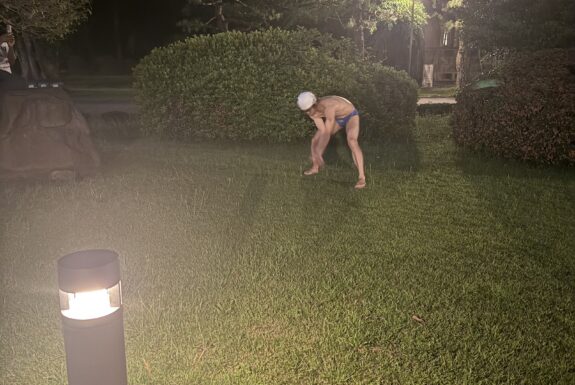 A Japanese man in a blue bathing suit, white swim cap and dark swim goggles is bent over, legs spread wide, on a grassy lawn. It is evening, and he is lit by outdoor lights. Behind him are bushes and trees.