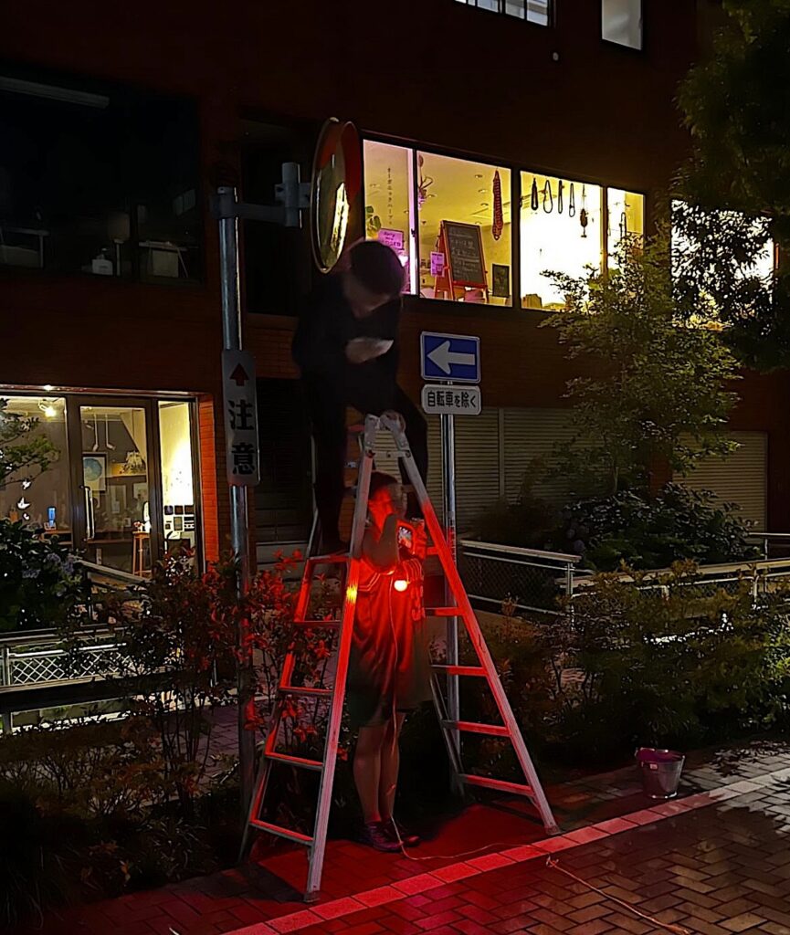 A man stand on top of a ladder at the edge of the street. He holds a white bowl in his hands. It is evening. A woman stands below the ladder bathed in a red light.