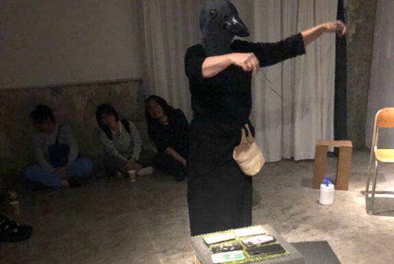 A human figure, dressed in black and wearing a crow's head, holds a string in front of them dangling it over an array of cellphones laid out on a box in front of them. A light coloured basket hangs from their waist.