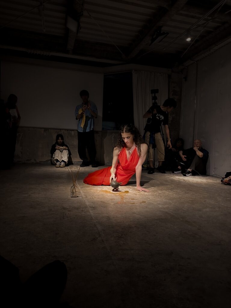 A figure in a full-length red dress sits on the concrete floor, propped up by one hand. In their other hand, they hold a Japanese-style teapot, pouring a dark liquid onto the floor. A glass jar with incense sticks sits beside them.