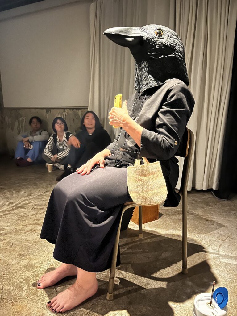 A barefoot human figure dressed in black with a crow's head sits in a chair. They hold a cellphone in their hand.