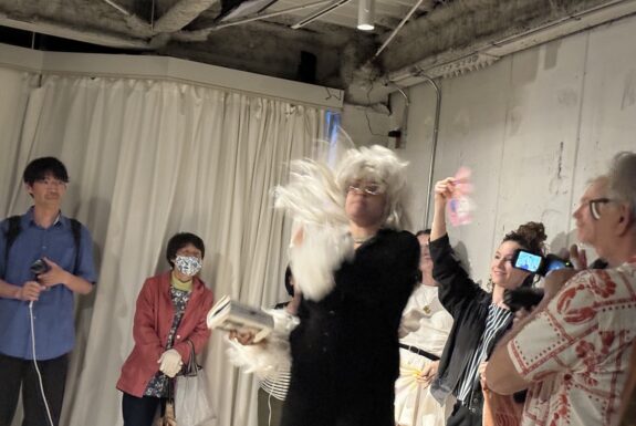 A woman dressed in black with a large white wig and sunglasses with gletter frames holds a photo book in her hand as a crowd looks on. She is being fanned by a woman who stands behind her.