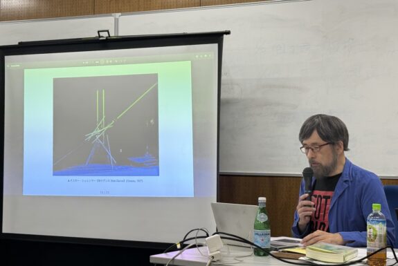 A white screen shows an image of Oskar Schlemmer's "Stick Dance.", Misafume Fukagawa sits at a table beside, looking at a laptop and speaking into a handheld microphone. He is bearded with glasses, and wearing a blue short unbuttoned, revealing a a black and red t-shirt with text.