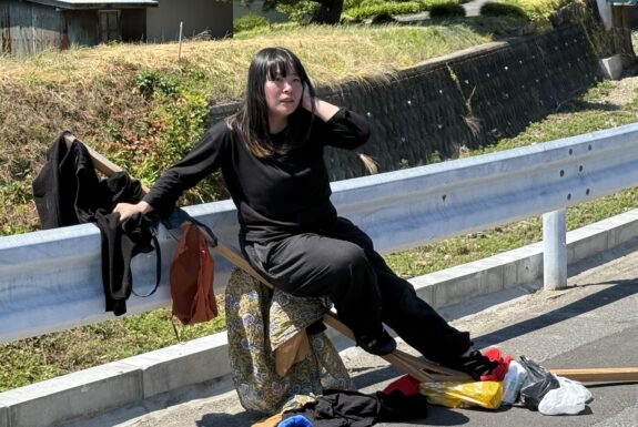 A woman dressed in a black top and black pants sits on a broken wooden board that is propped against a guardrail at the side of a road. She is holding one hand up to her ear, while the other supports her body against the guard rail. An assortment of clothes and bags hang from the board.
