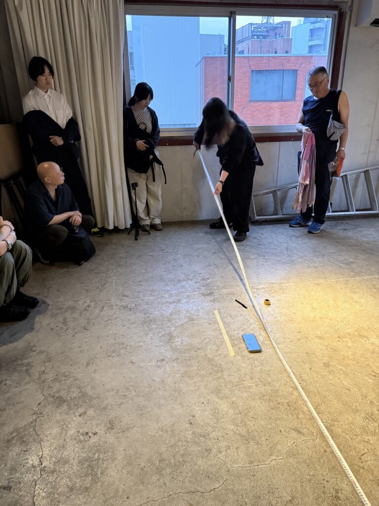 A woman dressed in black top and pants holds a measuring tape up against a wall. The tape extends forward and out of the frame. Some small implements, including a cellphone, a pen, and a roll of yellow tape sit on the concrete floor in front of her.
