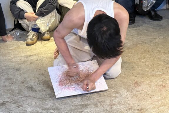 A man dressed in white pants and a white sleeveless t-shirt kneels on the floor. The image is shot from above, so we cannot see his face. He is drawing on a board with ballpoint pens in both hands. His forceful strokes etch into the board more than they leave traces of ink. His hand are moving quickly, so they are blurry.