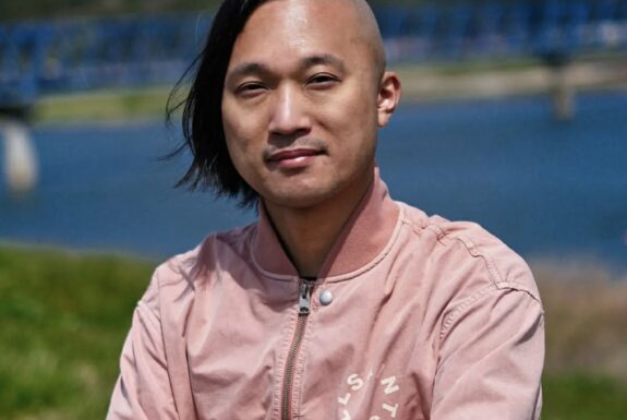 A man stands in front of a blurry outdoor backdropl He has long black hair on one side, shaved on the other. He wears a pink jacket. His arms are crossed.