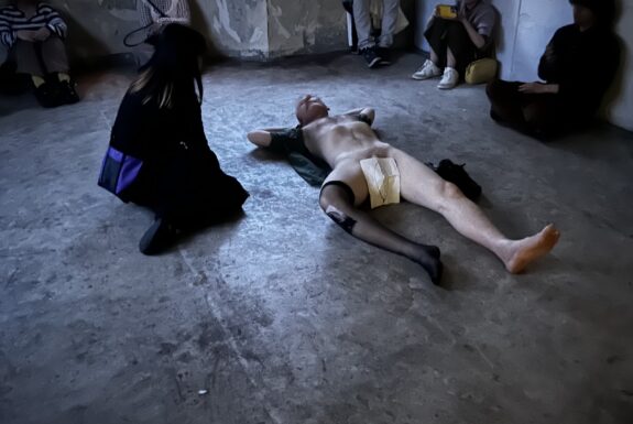 A naked man lies on a concrete floor. One leg is covered in a black stocking that comes to his thigh. The second stocking is draped over one arm. His genitals are covered by a paper drawing of a leaf. A woman dressed in black kneels beside him, looking into his face.