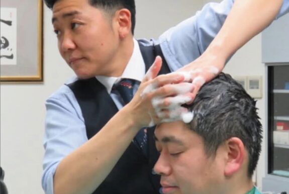 A barber in a light blue shirt with rolled-up sleeves, navy vest and tie washes the hair of a customer, who is covered in a green cape.