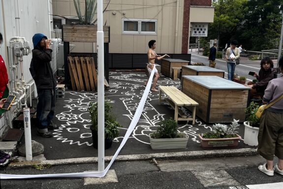 A male figure dressed in a white "fundoshi" (traditional Japanese underwear) that has a long cloth that trails for many metres and out of the frame of the camera stands in the middle of a tiny street corner parkette. His arms are raised in front of him, and one leg reaches back behind him.
