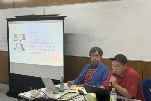A white screen shows a slide with Japanese text. Beside, two men sit at a table. Tomotsi, dressed in a red shirt and a baseball cap, looks at a laptop and speaks into a handheld microphone. Misafume Fukagawa, bearded with glasses, and wearing a blue short unbuttoned, revealing a a black and red t-shirt with text, gazes at Tomotsi as he speaks.