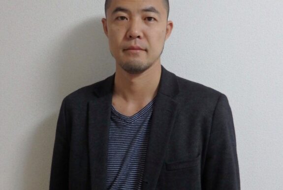 A man stands in front of a white backdrop. He wears a black blazer and a t-shirt with blue and white horizontal stripes.