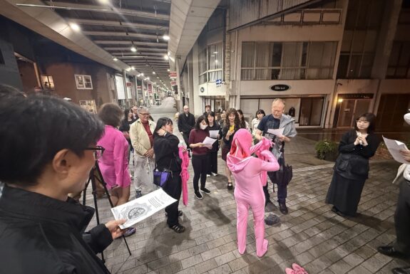 A figure wearing multiple pink zentai suits stands in the centre of a covered street, with several audience members surrounding them. Their back is to the camera. They are pulling another pink suit onto their arms like an additional skin. The audience members hold white papers with lyrics and a large graphic image in their hands.