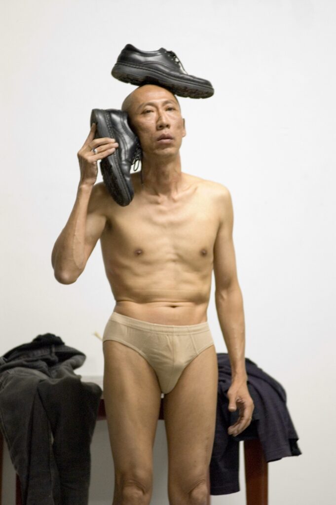 Lee Wen, an Asian man, stands almost naked, wearing only a pair of skin-coloured underwear. A black running shoe is balanced on his head. He holds a second black running shoe in his hand, held up to his ear like a telephone.