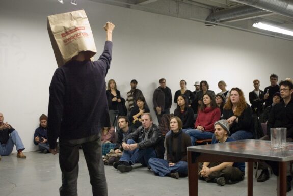 Lee Wen stands with his back to the camera, dressed in a black long-sleeve t-shirt and black pants. He has a brown paper shopping bag over his head. One fist is raised above his head, arm extended. A seated audience watches.